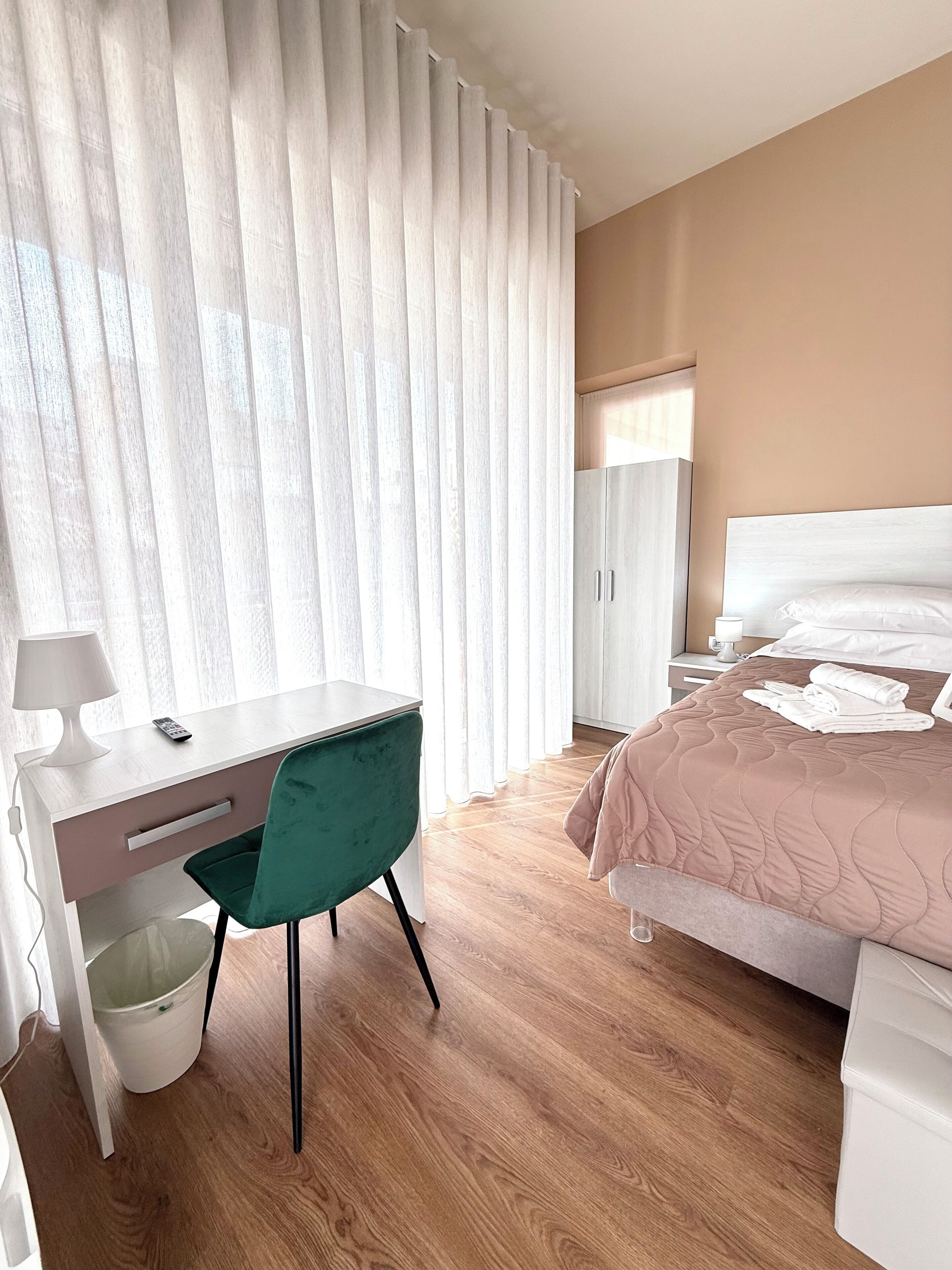 Una habitación de hotel moderna con suelo de madera, un escritorio con silla, una cama con colcha rosa y un armario.