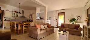 A living room with a sofa, a dining table with chairs, a kitchen area with cabinets, and a large window. - Villa gli Ulivi Calafate Lake View Varenna (Perledo)