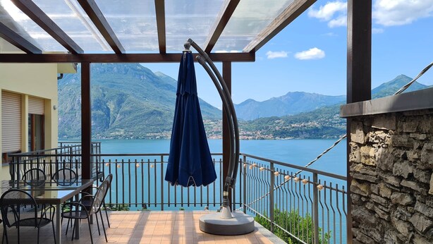 Un balcón con mesa y sillas, una sombrilla azul y vistas a un lago y montañas.