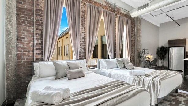 A hotel room with two beds, a kitchenette, and a view of a building through large windows. - Cozy Penthouse Apt w/Free Valet, Gym, Pool, View (Dallas)