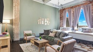A cozy living room with a sofa, armchairs, a coffee table, and a bookshelf. There is a large window with curtains and a view of the city. - Cozy Penthouse Apt w/Free Valet, Gym, Pool, View (Dallas)