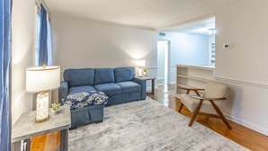 A living room with a blue sofa, a wooden chair, a small table with a lamp, and a bookshelf. - Suite B at Blue Sky Terrace (Columbus)