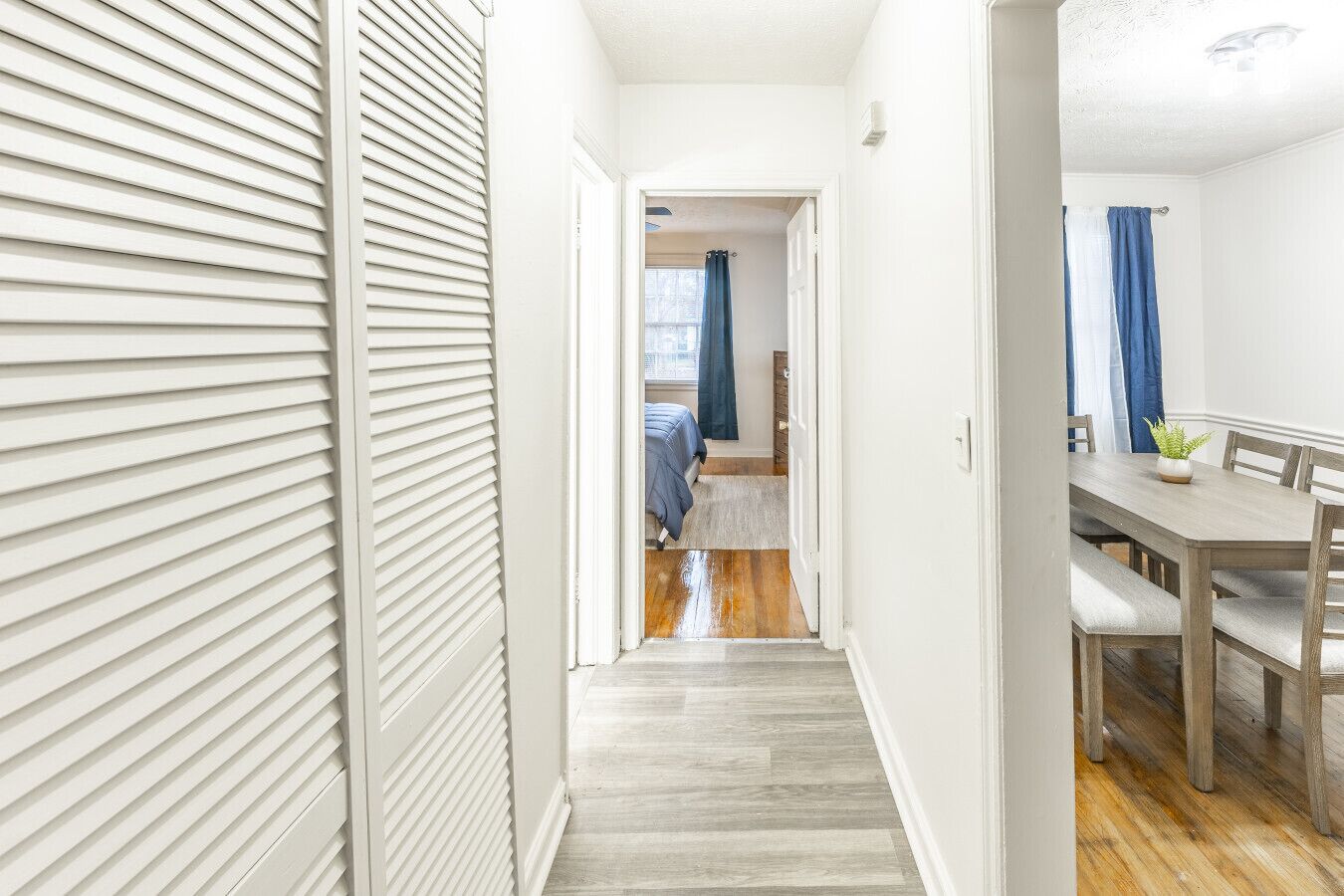 A hallway leading to a bedroom with a wooden floor, a dining area with a table and chairs, and a blue curtain.