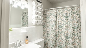 A bathroom with a white sink, a toilet, a shower curtain with a floral pattern, and a mirror with lights above it. - Suite B at Blue Sky Terrace (Columbus)