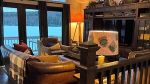 A cozy living room with a fireplace, a large window overlooking a snowy landscape, and a wooden railing. - Charming 2-bedroom lake house with AC in Parsonsfield (Parsonsfield)