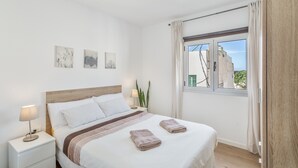 Een slaapkamer met een bed, nachtkastje, lamp, wanddecoratie, raam en gordijnen.