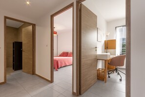 Ein modernes Hotelzimmer mit einer Holztür, einem Schreibtisch mit Stuhl, einem Bett mit roter Bettdecke und Blick ins Freie.
