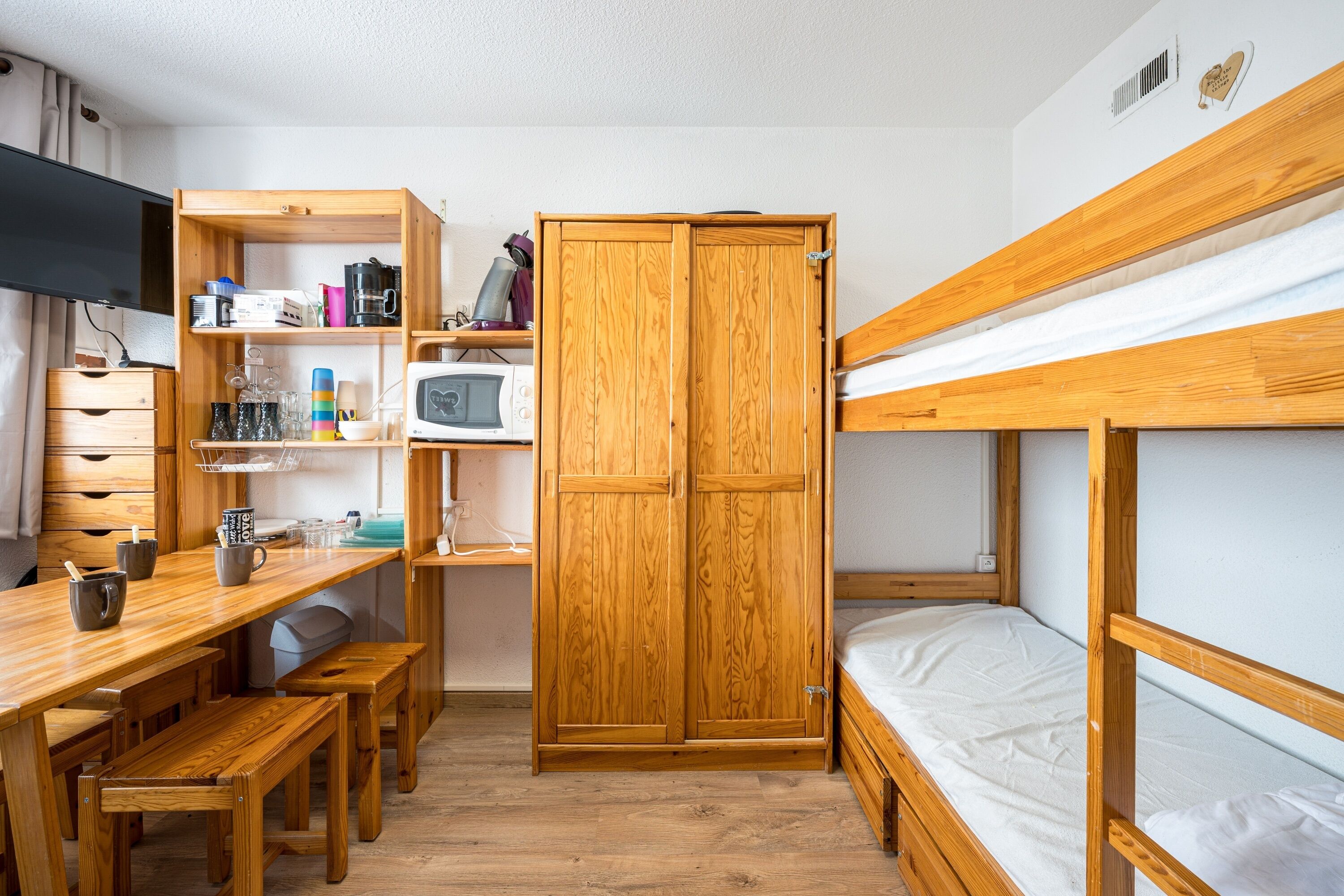 Ein kleines Zimmer mit Etagenbett, einem Holztisch mit Stuhl, einer kleinen Küchenzeile und einem Kleiderschrank.