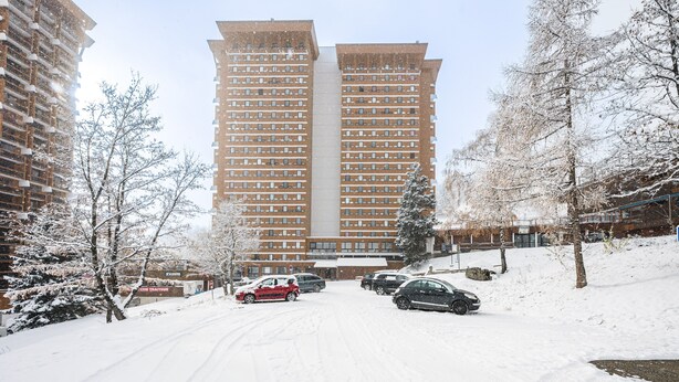 A snow-covered parking lot with cars and a tall building in the background. - Le Cocon des Sommets - Studio, station à pied (Villarembert)