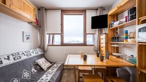 A cozy cabin with a wooden table, a sofa with a patterned blanket, a window with a view of mountains, and a television on a shelf. - Le Cocon des Sommets - Studio, station à pied (Villarembert)