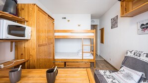 A compact room with a bunk bed, a microwave, a wooden cabinet, a table with a mug, and a couch with patterned cushions. - Le Cocon des Sommets - Studio, station à pied (Villarembert)