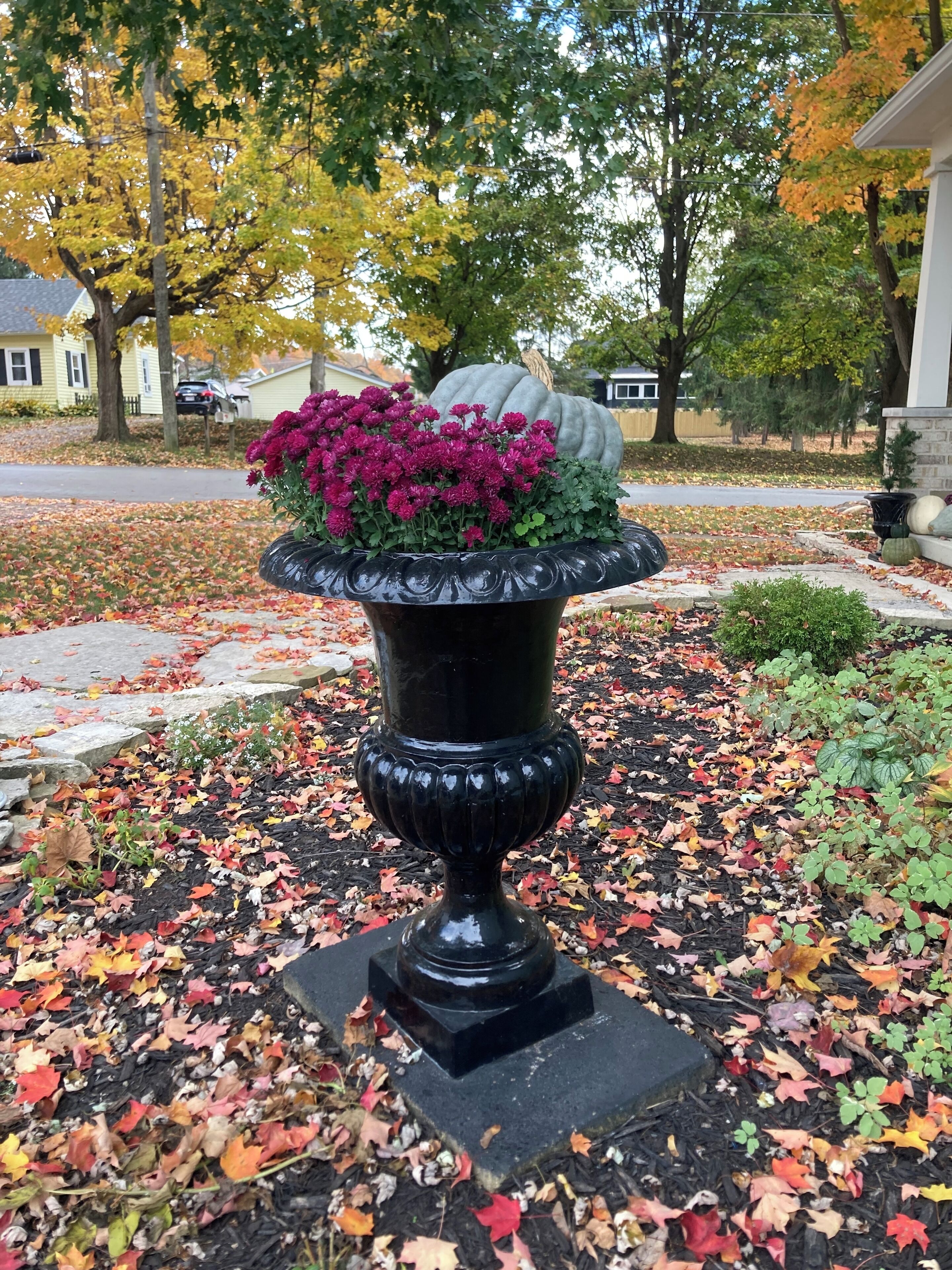 Eine schwarze Urne mit rosa Blumen und einem dekorativen Kürbis auf einem Steinsockel, umgeben von herabgefallenen Blättern.