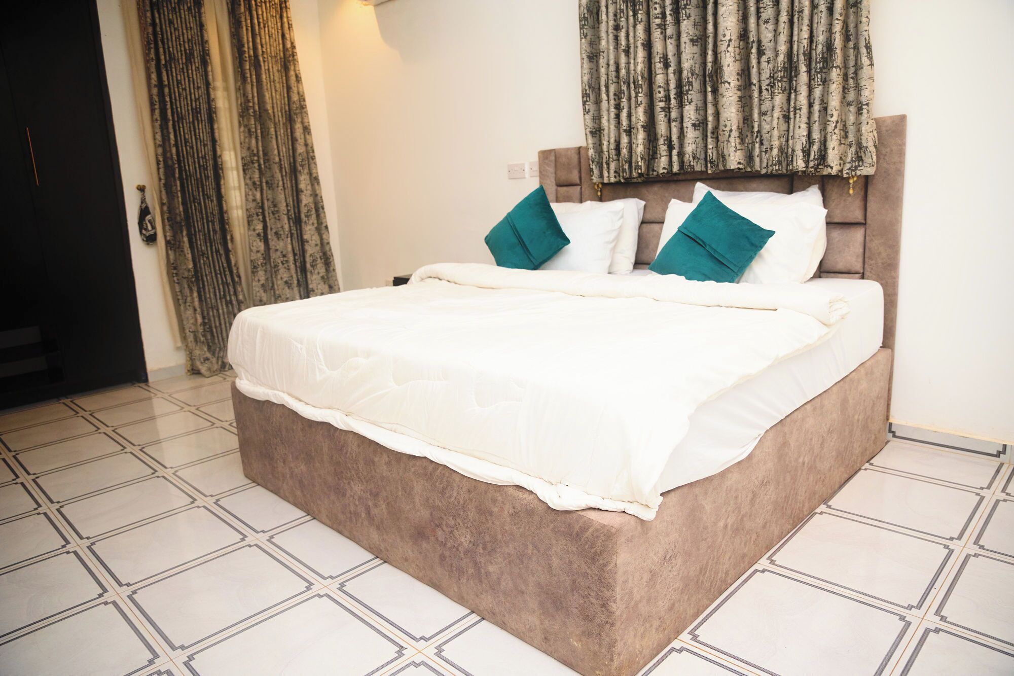 Cama bem arrumada com colcha branca e almofadas azul-turquesa, cabeceira marrom e cortina estampada à esquerda.