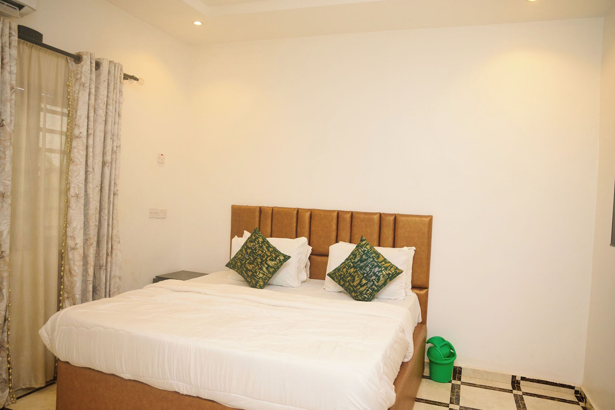 A neatly made bed with white bedding and two decorative pillows, a brown upholstered headboard, and a green bucket on the floor.