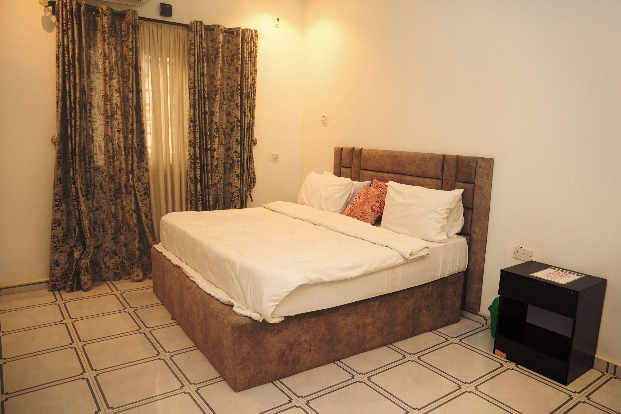 A neatly made bed with a wooden headboard, a pair of curtains, and a small black nightstand with a lamp.