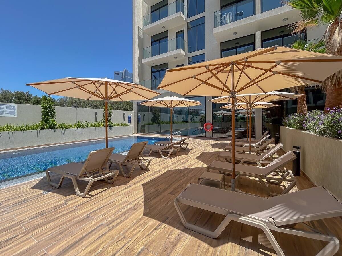 Un espace aménagé au bord de la piscine, avec des chaises longues et des parasols, une piscine à l’eau d’un bleu limpide et, en arrière-plan, un bâtiment moderne.