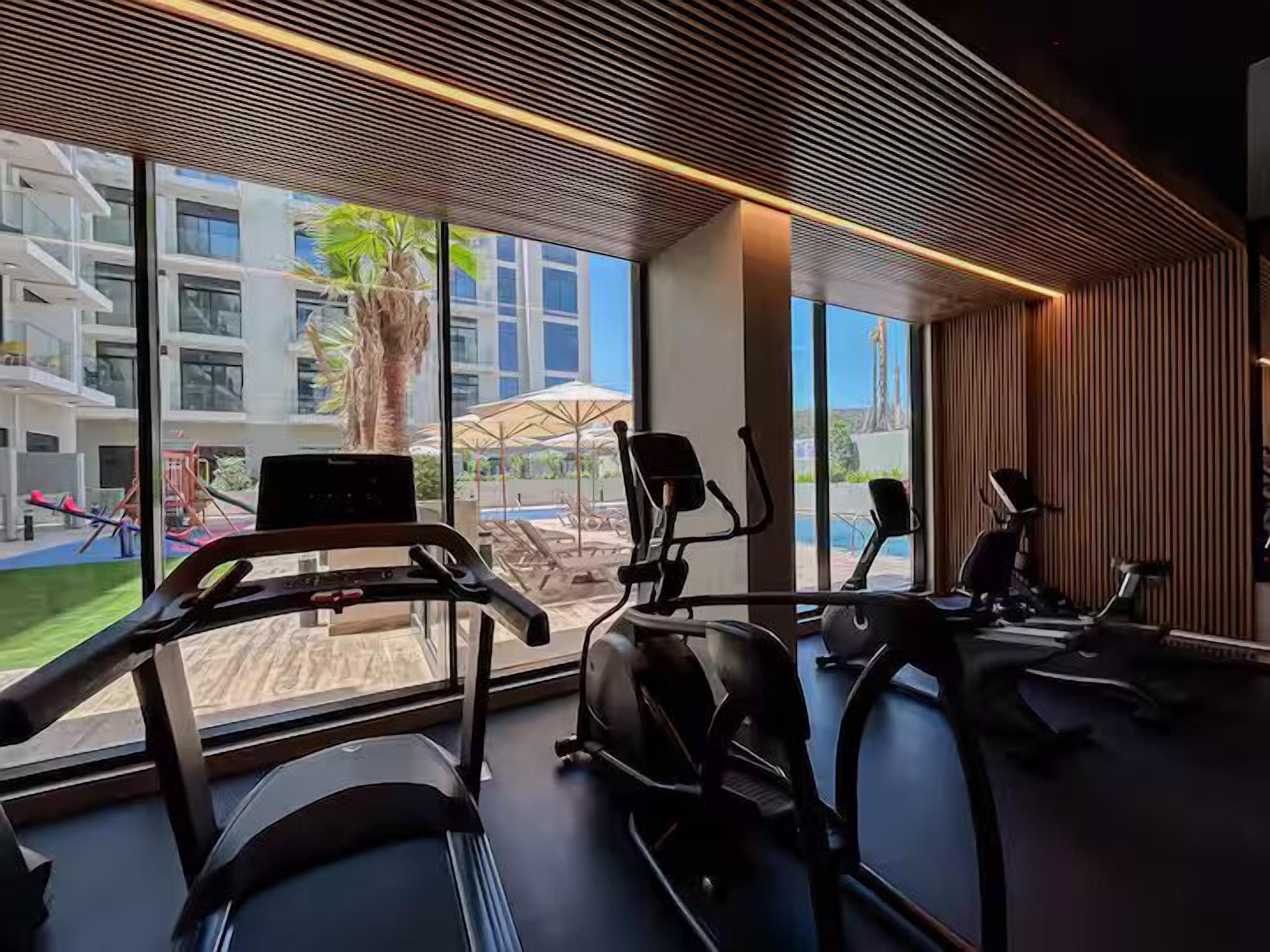 Un gimnasio con cintas de correr, bicicletas estáticas y máquinas elípticas, amplios ventanales que ofrecen vistas a una piscina y zona exterior.