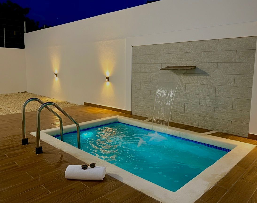A rectangular pool with a waterfall feature, surrounded by wooden decking and a white wall with a textured design.