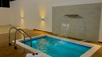 A rectangular pool with a waterfall feature, surrounded by wooden decking and a white wall with a textured design.