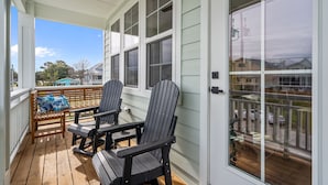 A covered porch with wooden flooring, two Adirondack chairs, a bench with a blue pillow, and a view of a neighborhood through large windows. - Island Sunrise Town Home 3 Bdrm/2.5 Bath Sleeps 12!   7 Min walk to the Beach! (Carolina Beach)