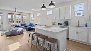 A modern kitchen with white cabinets, a central island with stools, pendant lighting, and a view of the living area with a blue sofa and a bean bag chair. - Island Sunrise Town Home 3 Bdrm/2.5 Bath Sleeps 12!   7 Min walk to the Beach! (Carolina Beach)