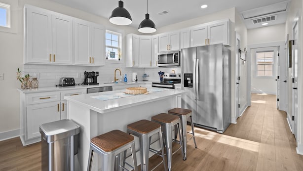 A modern kitchen with white cabinets, stainless steel appliances, and wooden stools. - Island Sunrise Town Home 3 Bdrm/2.5 Bath Sleeps 12!   7 Min walk to the Beach! (Carolina Beach)