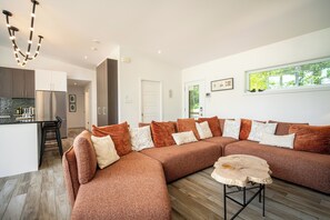 A modern living room with a large sectional sofa, a small round table, and a kitchen area with a refrigerator and cabinets.