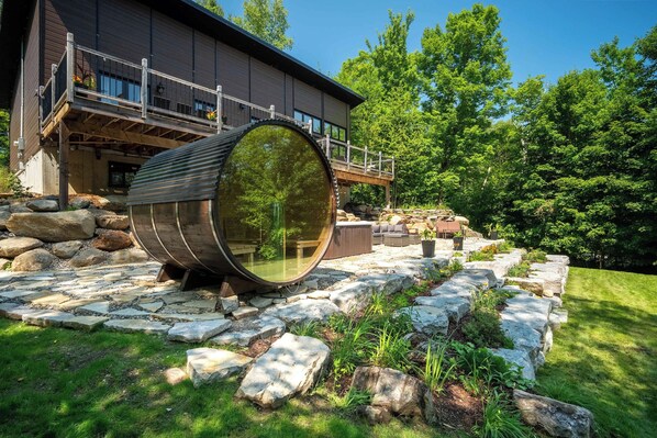 A modern building with a large cylindrical structure in the foreground, a wooden deck, and a stone pathway leading to a seating area.