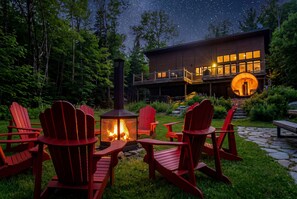 A modern lodge with a fire pit, red Adirondack chairs, and a deck overlooking a forest.