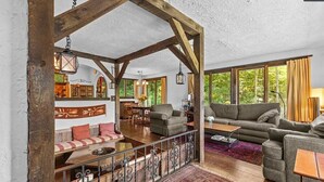 En rummelig opholdsstue med højt til loftet, træbjælker og store vinduer. Der er et siddeområde med sofaer og et sofabord.