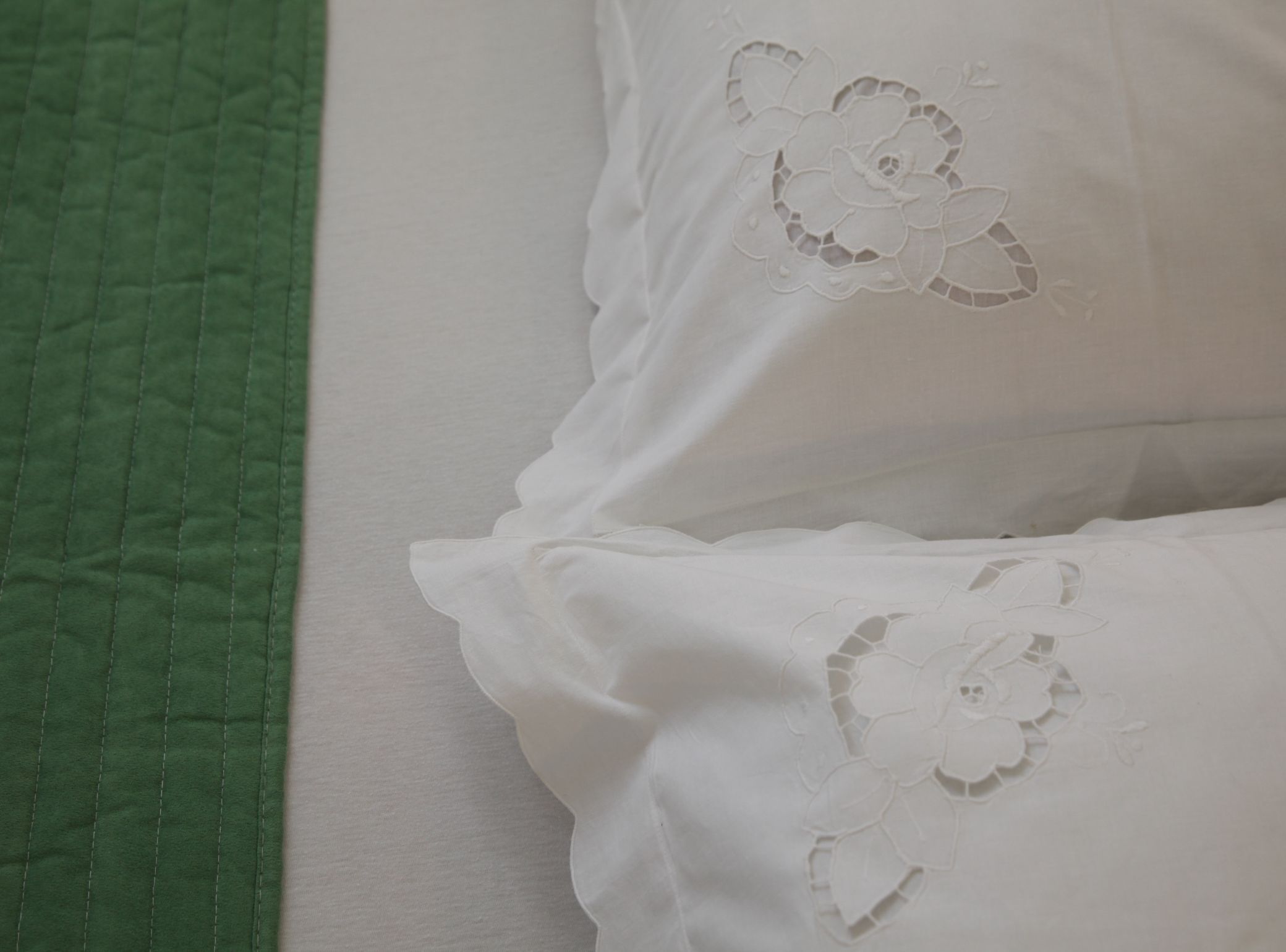 Una cama con colcha verde y almohadas blancas con bordado floral.