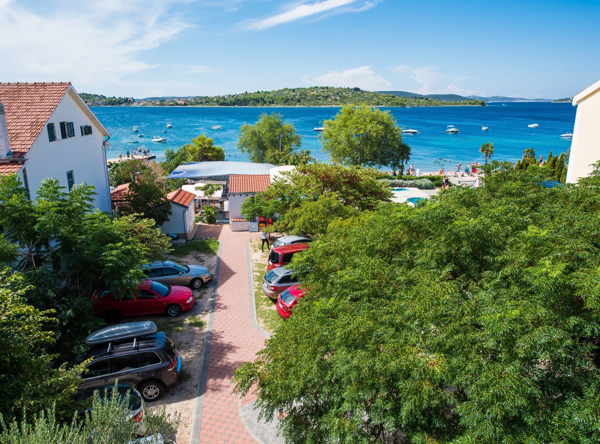 A coastal view with a paved pathway leading to a building with a red roof, parked cars, and a clear blue sea with boats.