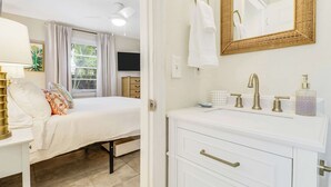 Habitación de hotel con cama, mesita de noche, lámpara, televisor y baño con lavabo, espejo y toallero.