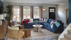 A living room with a blue sectional sofa, a round coffee table, and a wicker chair. There is a large window with curtains, a door, and a wall-mounted light fixture. - Pine & Paddle Getaway • Game Room • Hot Tub • Pool (Muskegon)
