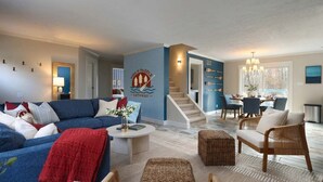 A modern living room with a blue sofa, a coffee table, and a wicker chair. There is a staircase leading to another level. - Pine & Paddle Getaway • Game Room • Hot Tub • Pool (Muskegon)