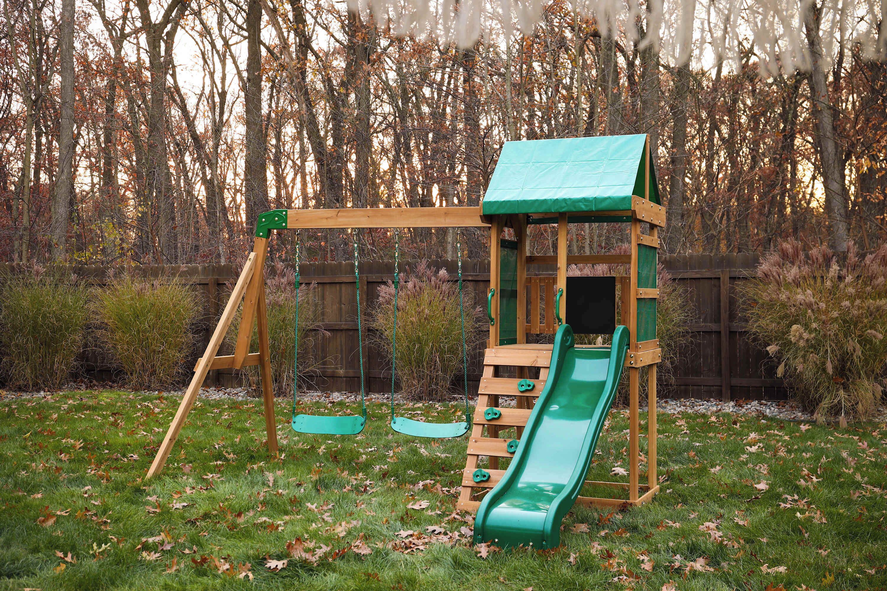 A wooden swing set with a green slide in a backyard.