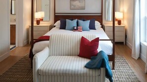 A four-poster bed with a striped upholstered chair, a nightstand with a lamp, and a large window with curtains. - Pine & Paddle Getaway • Game Room • Hot Tub • Pool (Muskegon)