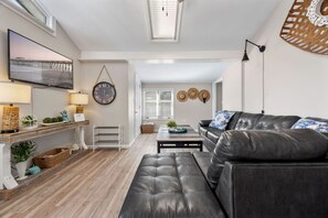 Ein modernes Wohnzimmer mit einer Ledersofa, einem Couchtisch und einem an der Wand montierten Fernseher. Ein Fenster mit Jalousien, eine Pendelleuchte und eine kleine Pflanze auf dem Couchtisch sind ebenfalls vorhanden.