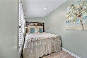 Un dormitorio con una cama, cabecera, cuadro de una palmera y una ventana con persianas.