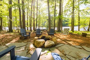 A campsite with a fire pit, several Adirondack chairs, and a view of a lake surrounded by trees. - Pet Friendly,Lakefront,Hot Tub,Close to State Park (Swanton)