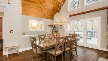 A dining area with a wooden table and chairs, a chandelier, and large windows with a view of trees.