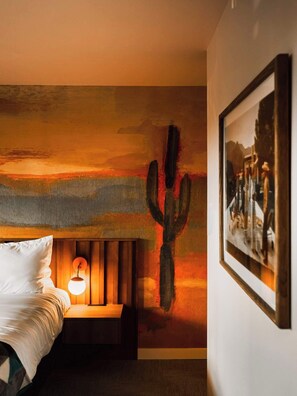 A bedroom with a bed, a wall-mounted lamp, a cactus painting, and a framed picture. - Prickly Pear Paradise | La Vista Motel | 204 (Denver)