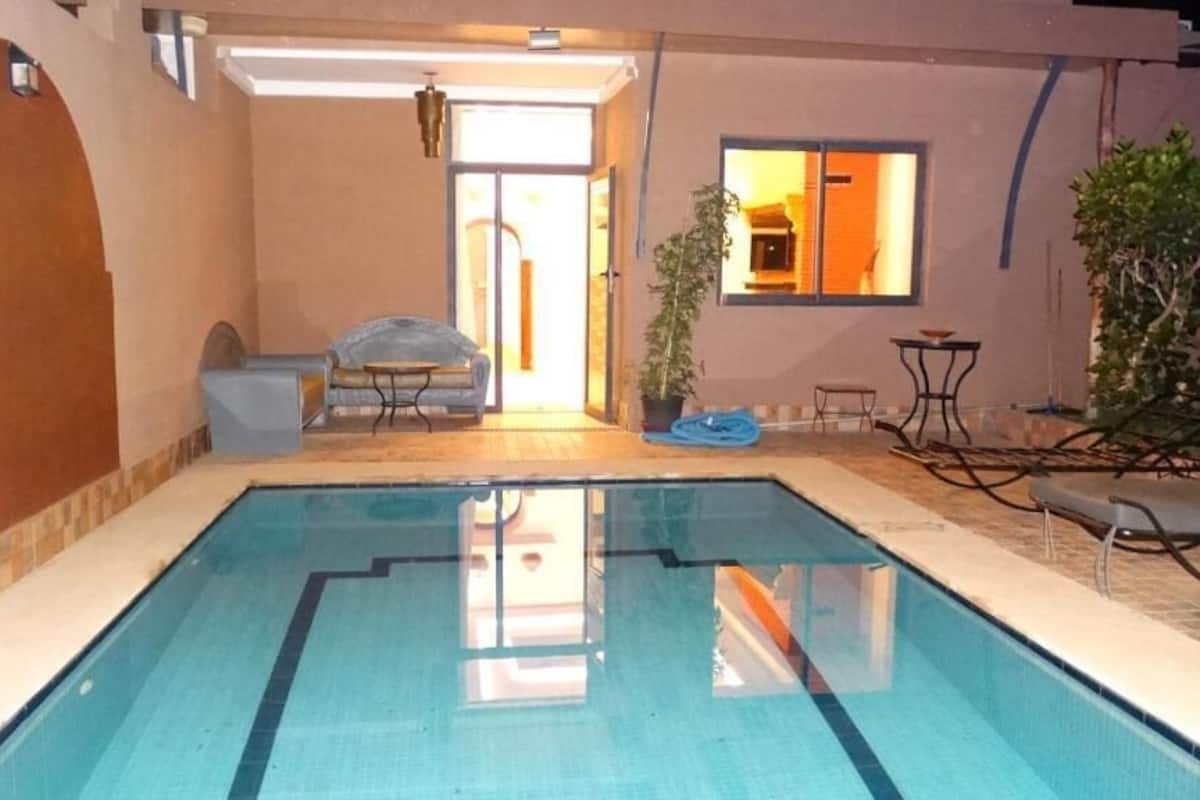 Une piscine dont le bord est clairement délimité par une ligne bleue, un espace détente couvert avec une table et des chaises, et une plante en pot.