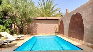 A swimming pool with a tiled deck, surrounded by a low wall and palm trees. - Villa des roses privée (Marrakech)