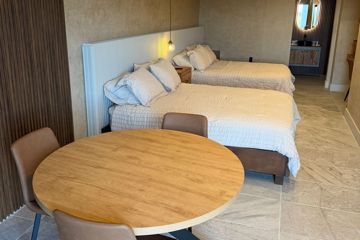A modern hotel room with two beds, a round wooden table, and a seating area.