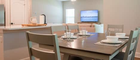 Un comedor con una mesa preparada para cuatro, un televisor al fondo y una cocina con refrigerador y fregadero.
