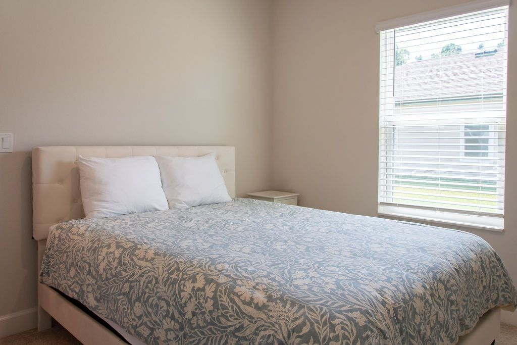 A bedroom with a bed, two pillows, a nightstand, and a window with blinds.