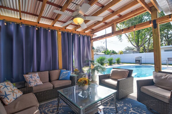 A covered outdoor seating area with a glass coffee table, wicker furniture, and a view of a backyard swimming pool. - Enjoy 3-Bedroom House with AC, Heated Pool and Gazebo in Charming Pinellas Park (Pinellas Park)