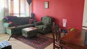 A living room with a red wall, a patterned rug, a wooden dining table, and a green armchair. - Colorful 1 bedroom Flat in South East London area (Bromley)