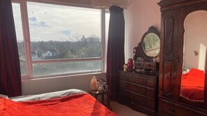 A bedroom with a bed, a dresser, a mirror, and a window with a view of houses and trees. - Colorful 1 bedroom Flat in South East London area (Bromley)